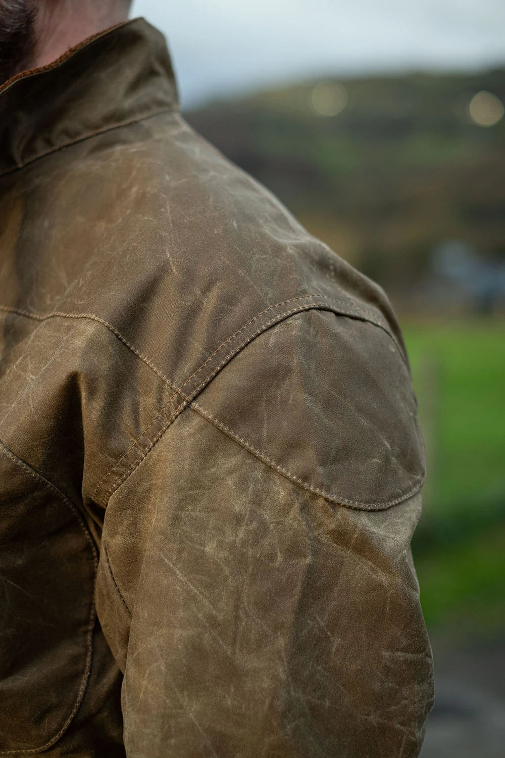 Close-up of a person wearing a brown waxed canvas jacket outdoors, showing the shoulder and upper back with a blurred natural background.