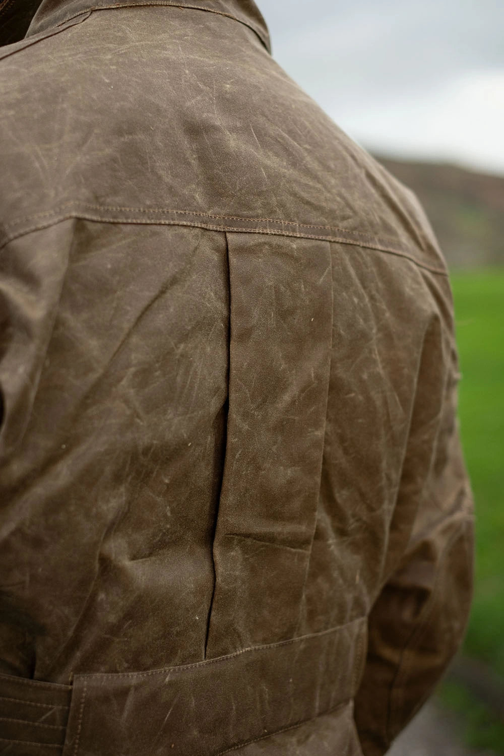 Close-up of the back of a brown waxed canvas jacket, showing the texture and stitching, with an outdoor blurred background.