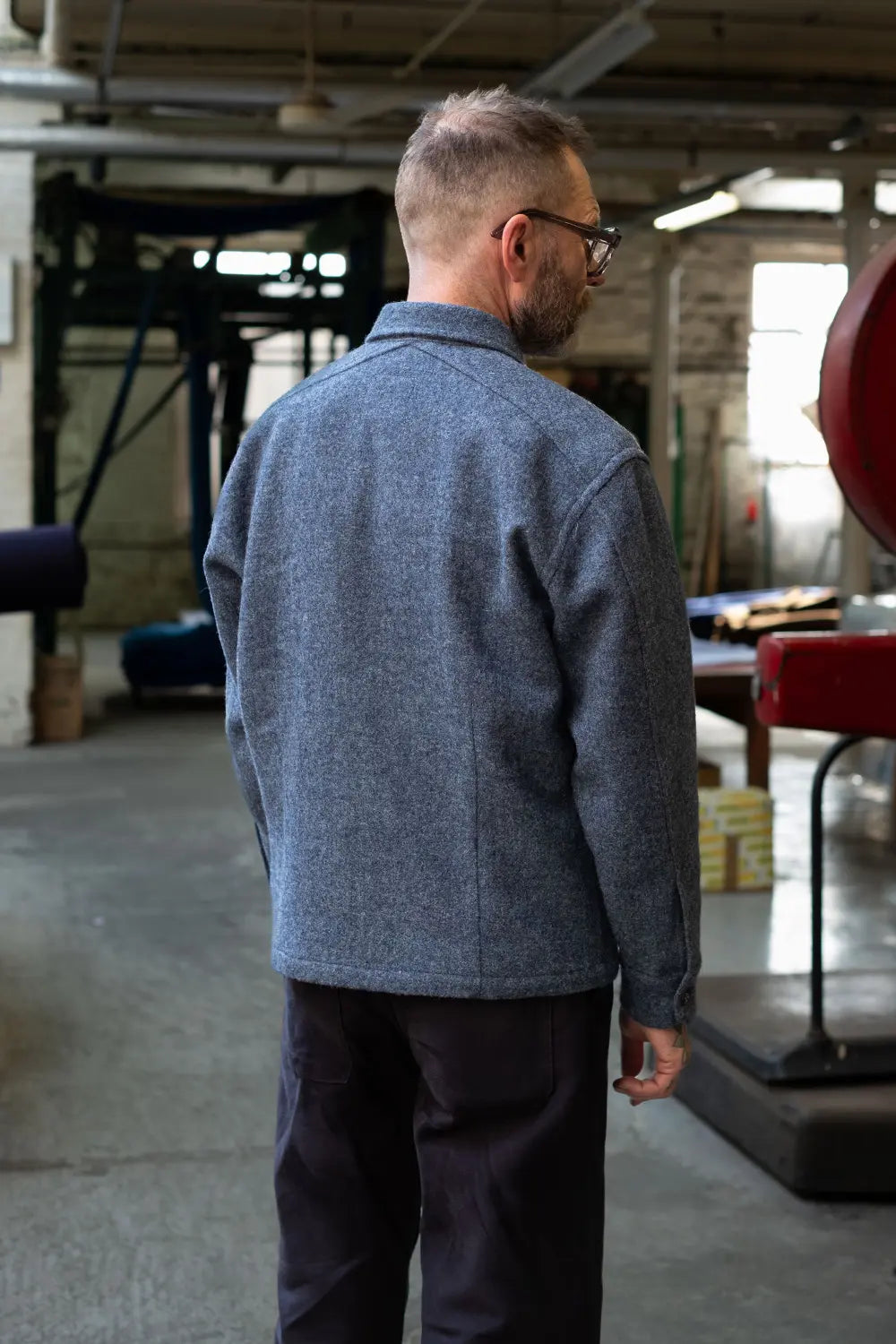 A man wearing a textured gray heathered button-up shirt and dark trousers, viewed from behind in an industrial workshop setting.