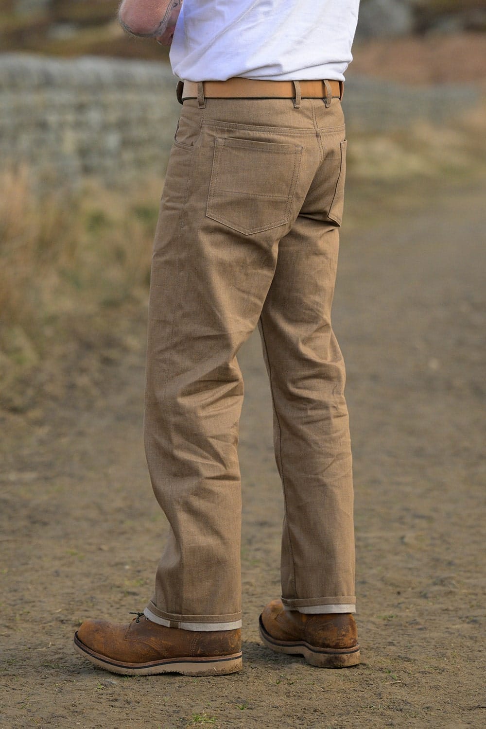 A person shown from the waist down wearing tan canvas work pants, a white t-shirt, and brown work boots, standing outdoors on a dirt path.