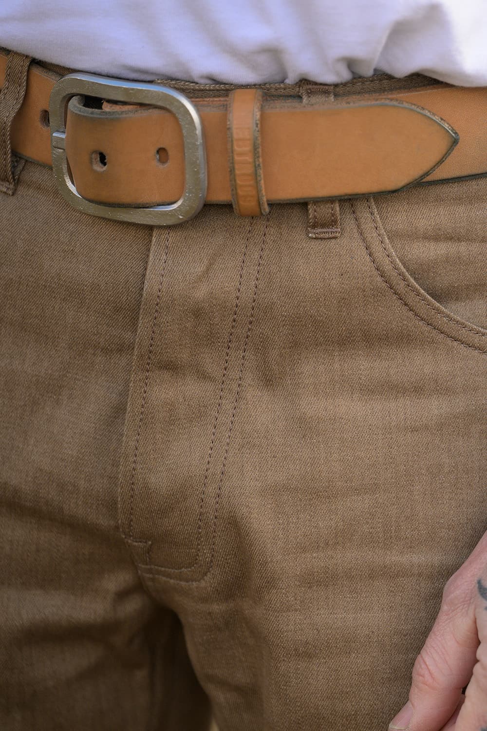 Close-up of a person wearing tan canvas pants with a light brown leather belt featuring a silver-toned metal buckle, paired with a white shirt.