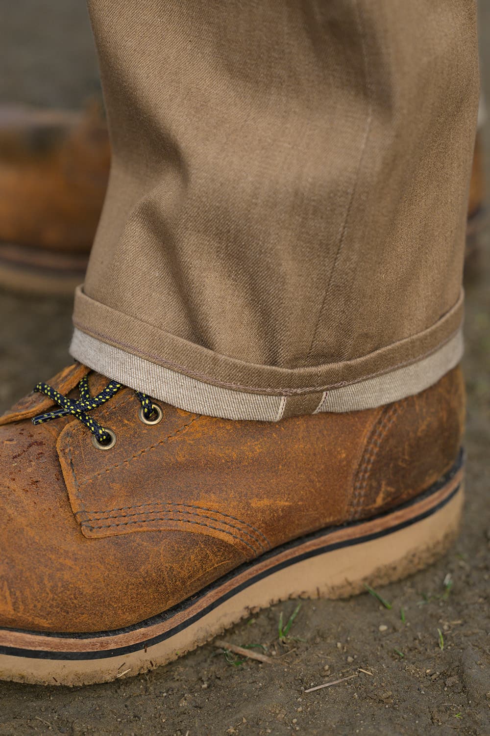 A close-up of a person's foot wearing a brown distressed leather lace-up work boot with tan rolled-up trousers, standing on dirt ground.
