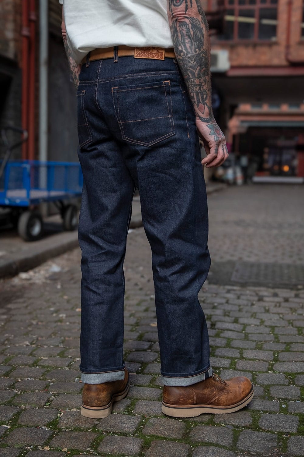 A person shown from the back wearing dark indigo denim jeans with a brown leather belt and rolled cuffs, standing on a cobblestone street.