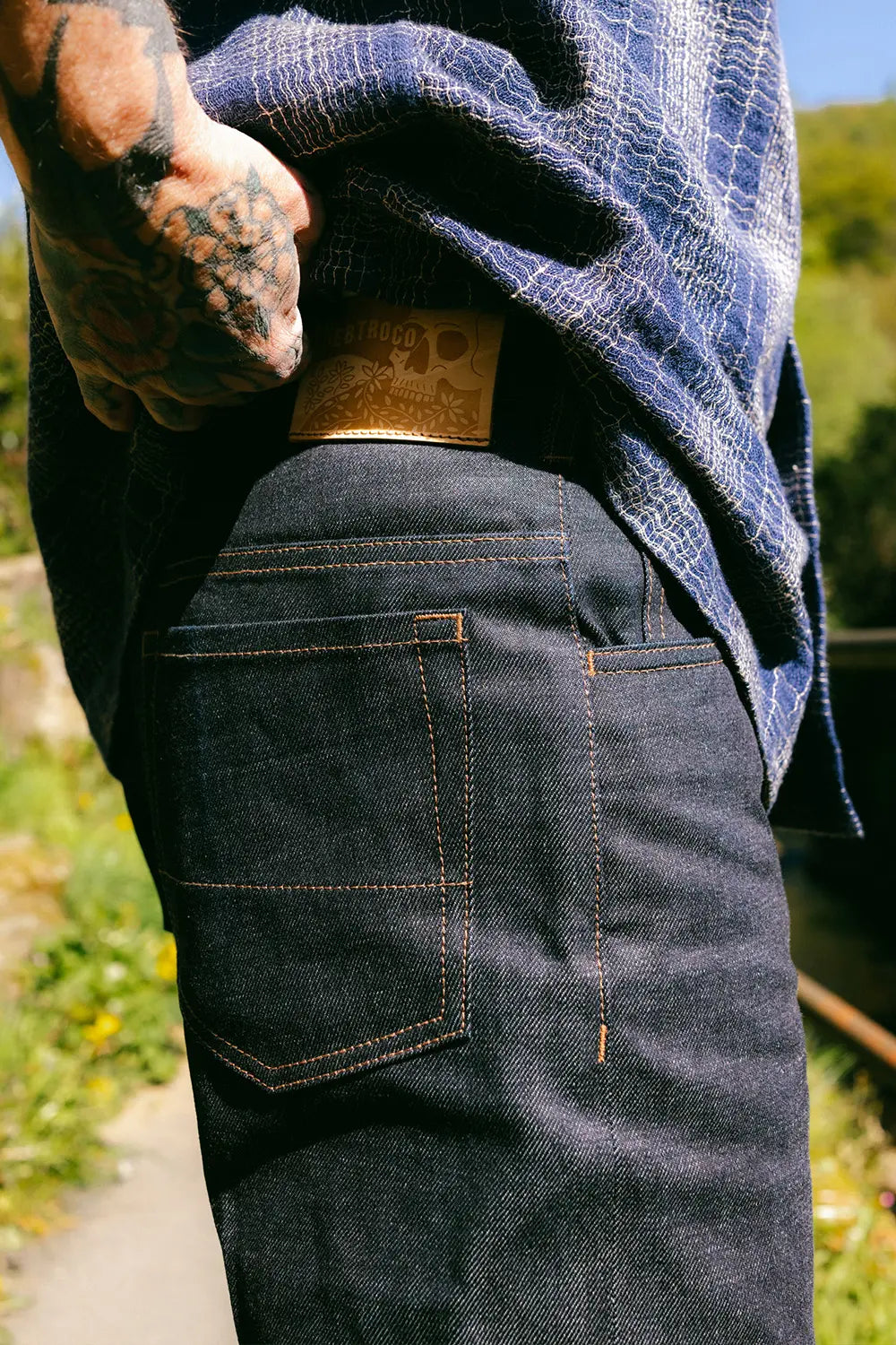 Close-up of a person wearing dark indigo denim jeans with contrast orange stitching, pulling up a blue plaid shirt to reveal a tan leather waistband patch. The background is a blurry outdoor green landscape.