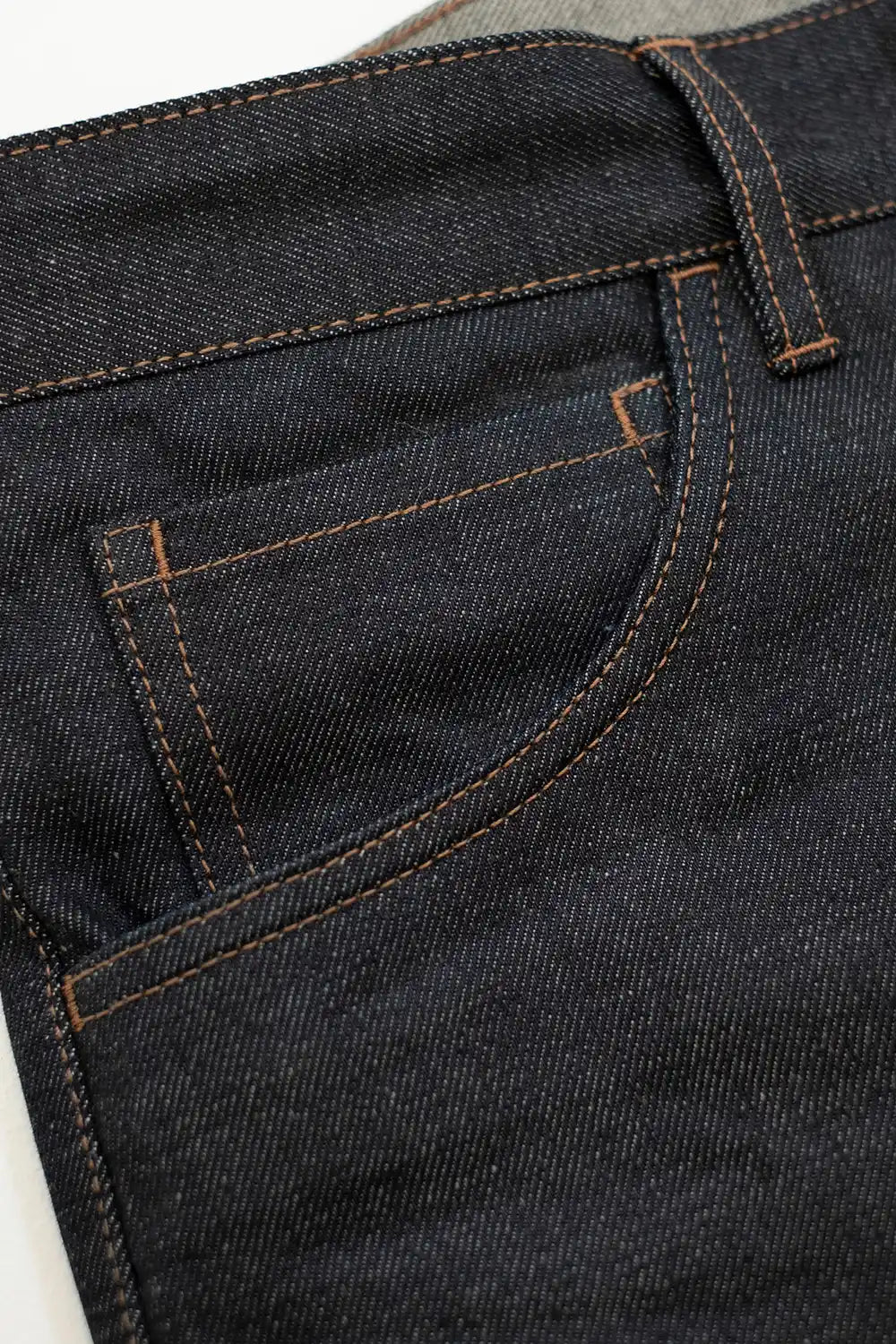 Close-up of dark indigo denim jeans showing the back pocket and belt loop with contrasting brown stitching.
