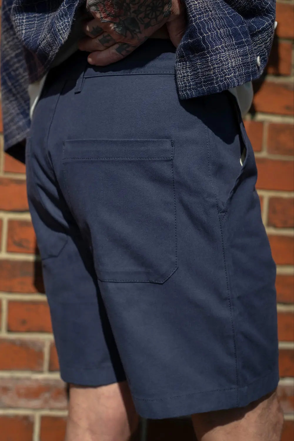 A close-up of a person wearing navy blue chino shorts, with a blue and white plaid shirt tucked into the waist and a brick wall in the background.
