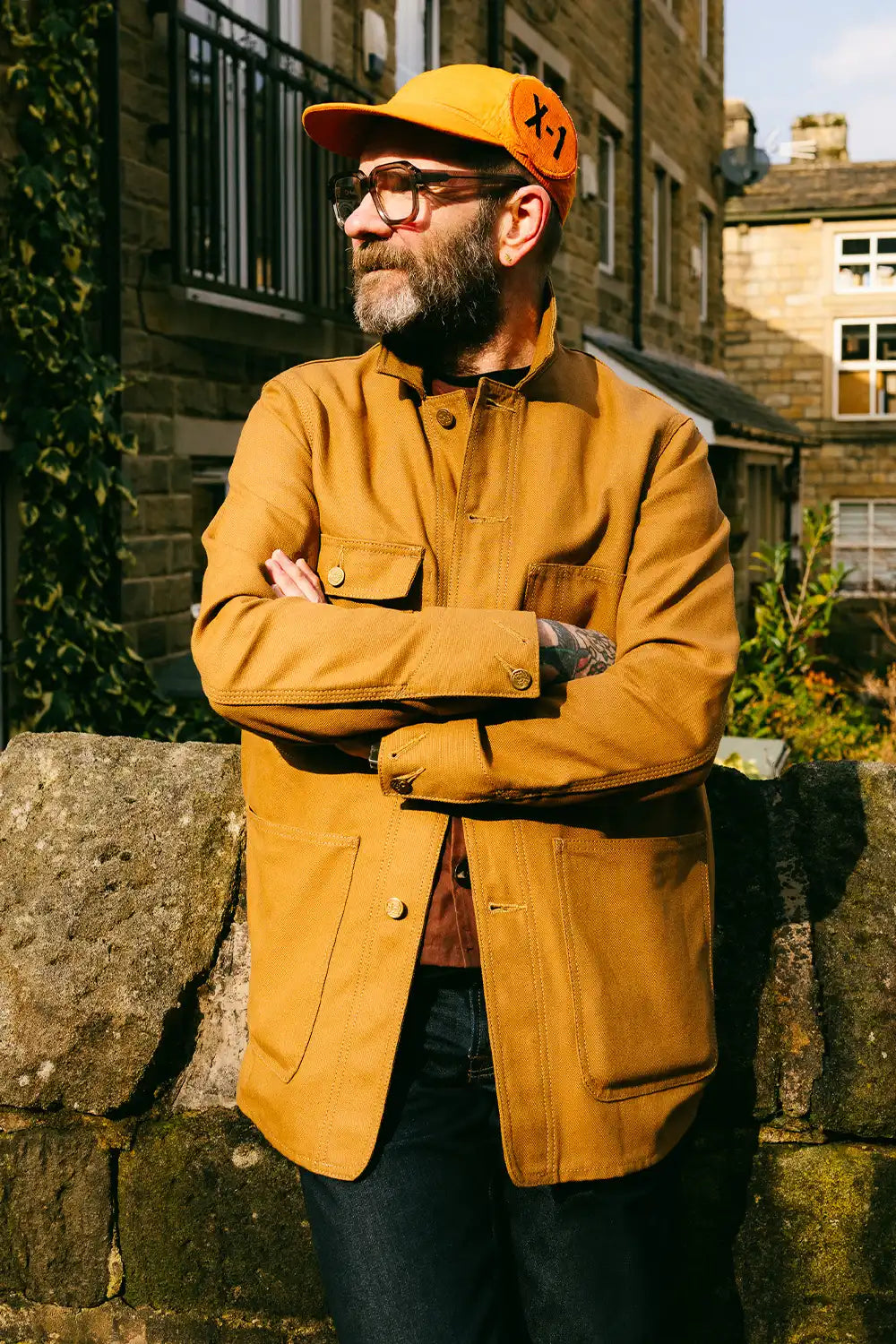 A man wearing a tan bob job chore coat, matching cap, and glasses, standing outdoors with arms crossed in front of a stone wall.