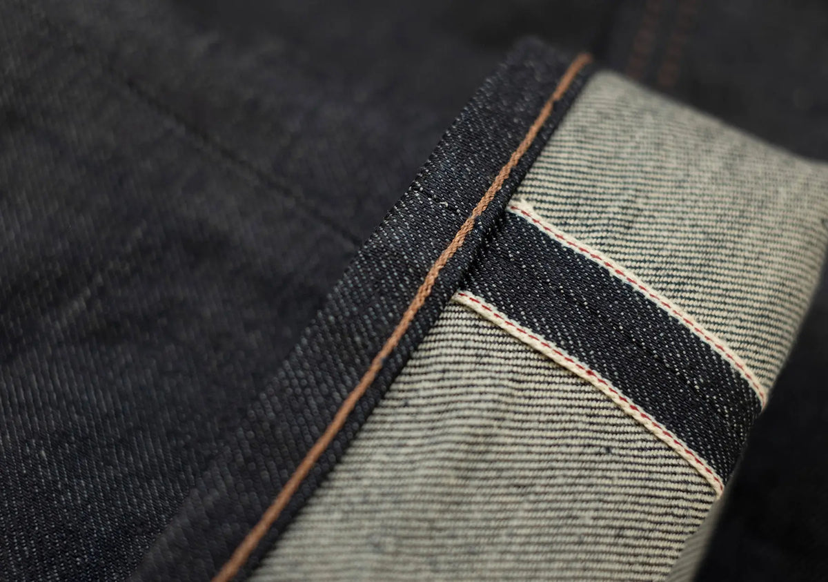 Close-up of dark indigo denim fabric with a folded hem showing the lighter gray ribbed inner side and contrast tan stitching.