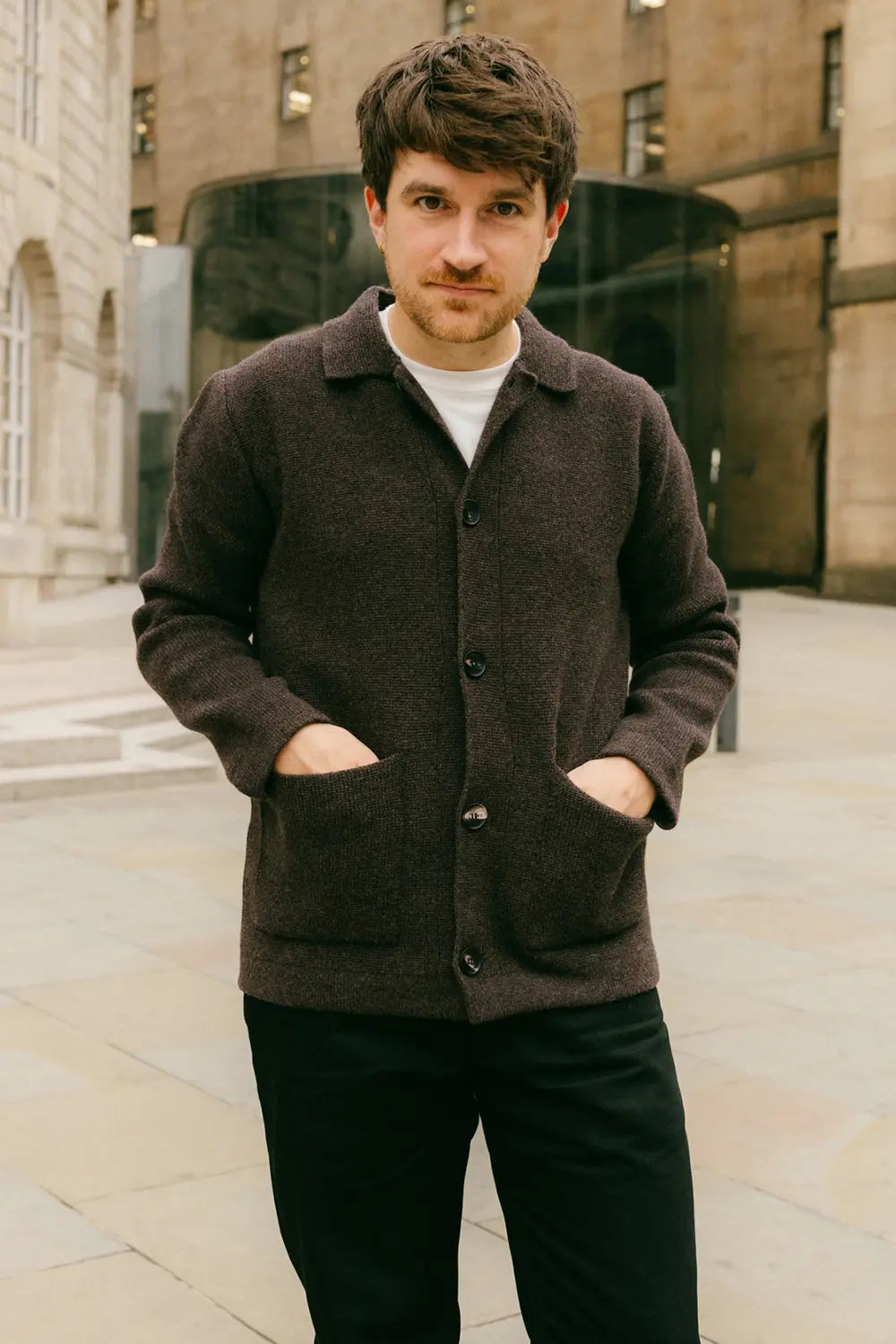 A man wearing a dark brown wool-blend button-up cardigan with front pockets over a white t-shirt and black trousers, standing outdoors in front of a stone building.