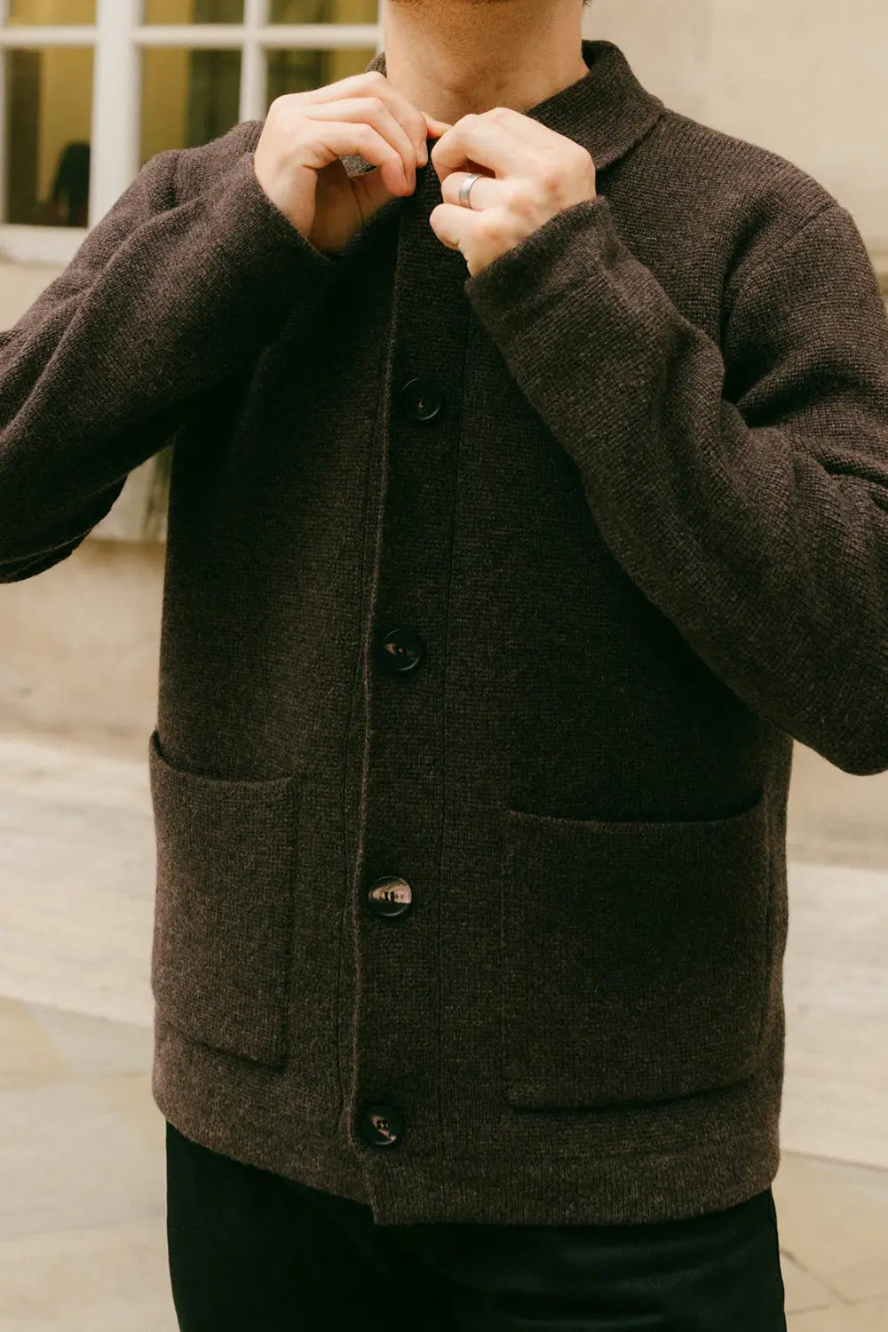 A person wearing a dark brown textured knit button-up cardigan with front pockets, shown from the neck down against an outdoor background.