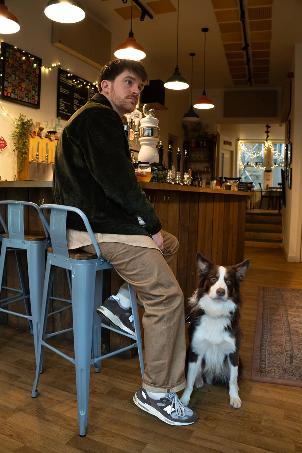 A man wearing a c major jacket, beige pants, and gray sneakers sits on a blue metal bar stool in a cozy bar or cafe, looking at a black and white dog sitting on the floor nearby.