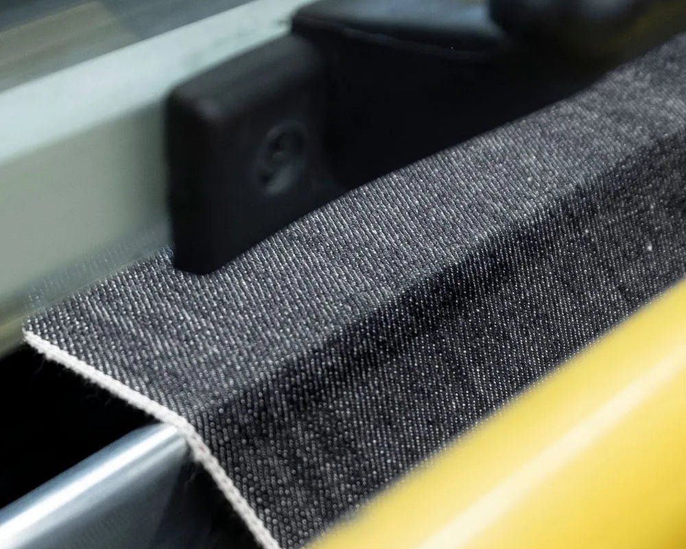 A close-up of a dark gray woven fabric strip with white edging, shown alongside a bright yellow cylindrical rod and black hardware.