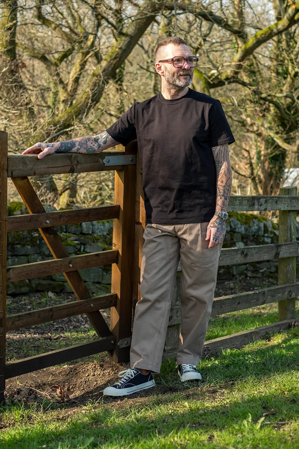 A man with tattoos and glasses stands outdoors next to a wooden gate, wearing a black t-shirt, beige trousers, and black canvas sneakers.