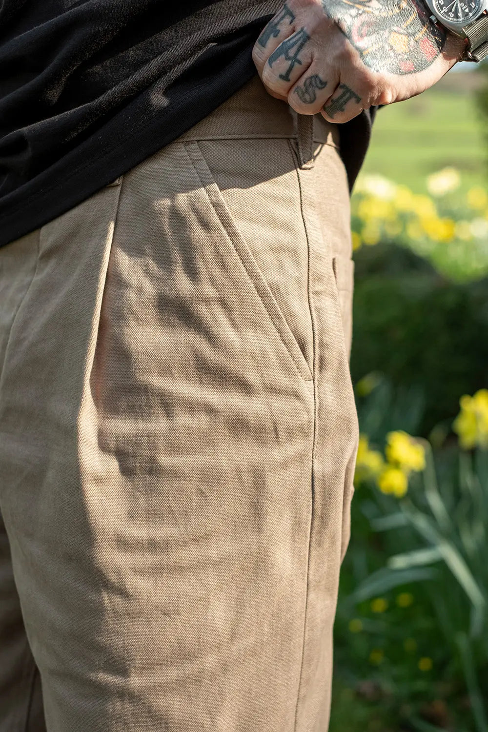 Close-up of a person wearing beige linen trousers and a black shirt, standing outdoors in a garden with yellow flowers blurred in the background.