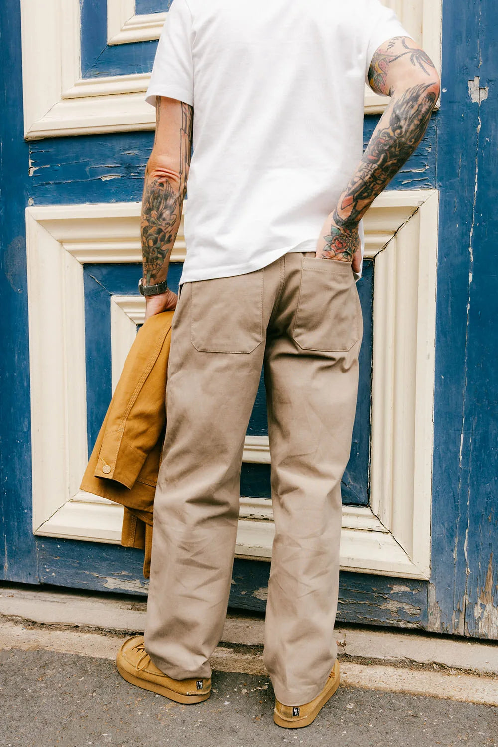 A man from behind wearing beige cargo pants, a white t-shirt, and tan shoes, holding a brown jacket against a blue wooden door.