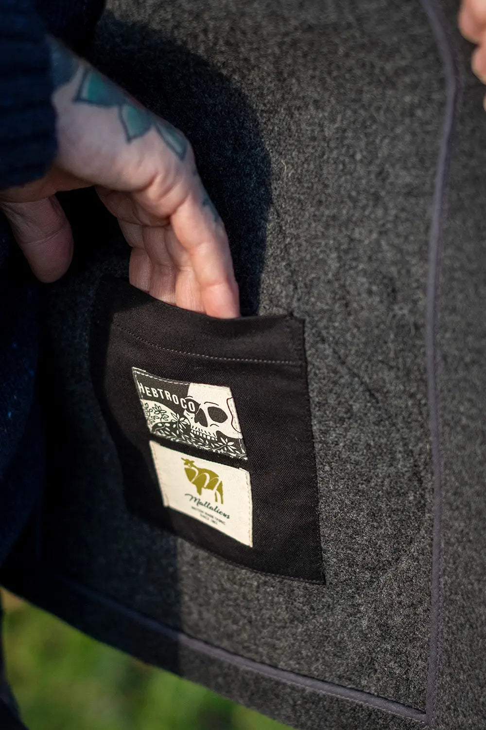 Close-up of a person's hand pulling open a black pocket on a dark gray textured jacket, revealing two brand labels: 'HERITAGE CO.' and 'The Northern'.