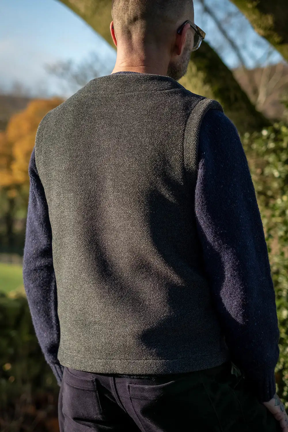 A man viewed from behind wearing a teasel duffel vest over a navy blue long-sleeve jumper, standing outdoors in a sunny garden setting.