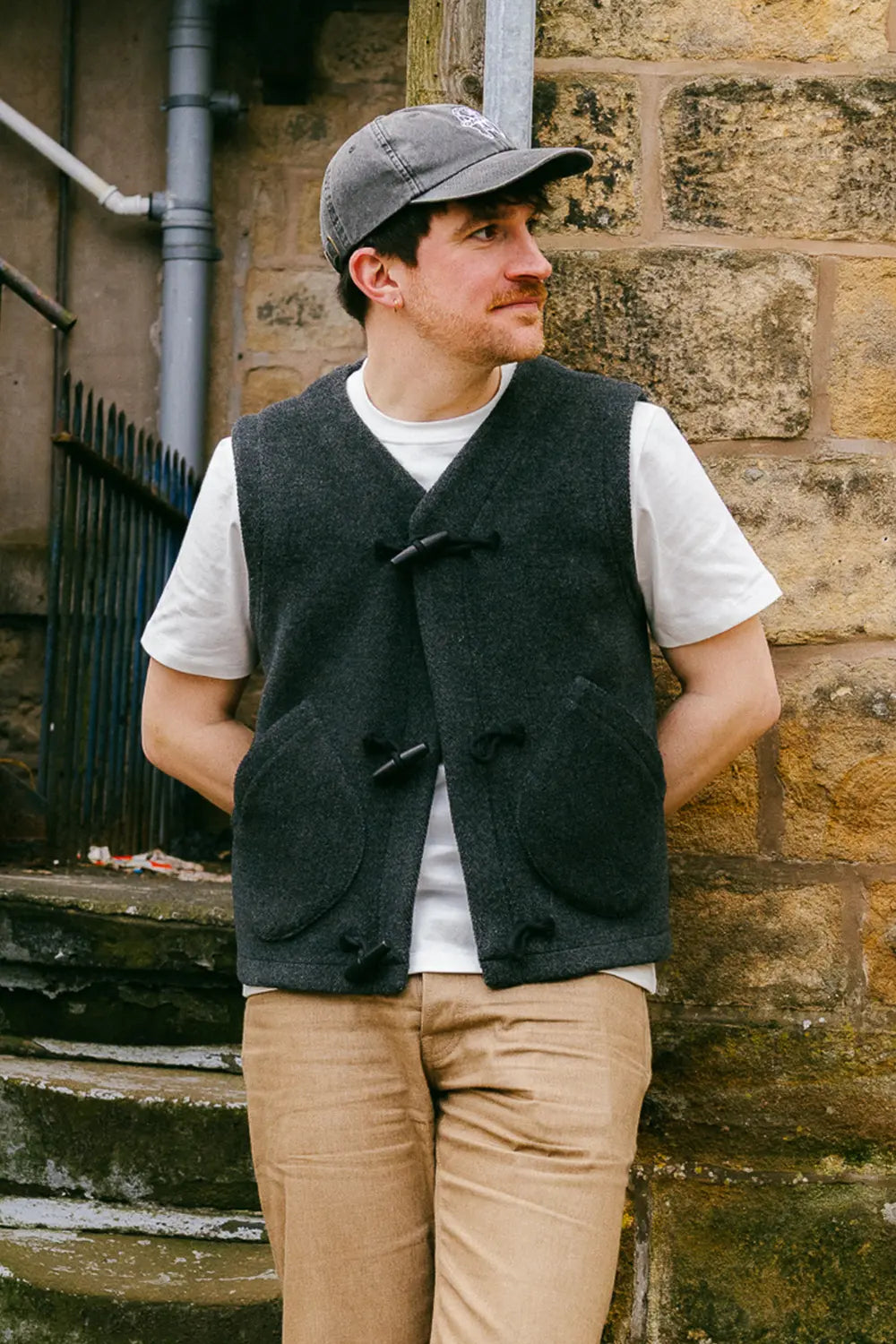 A man wearing a dark charcoal gray wool duffle vest over a white t-shirt, khaki trousers, and a gray baseball cap, standing against a rustic stone wall.