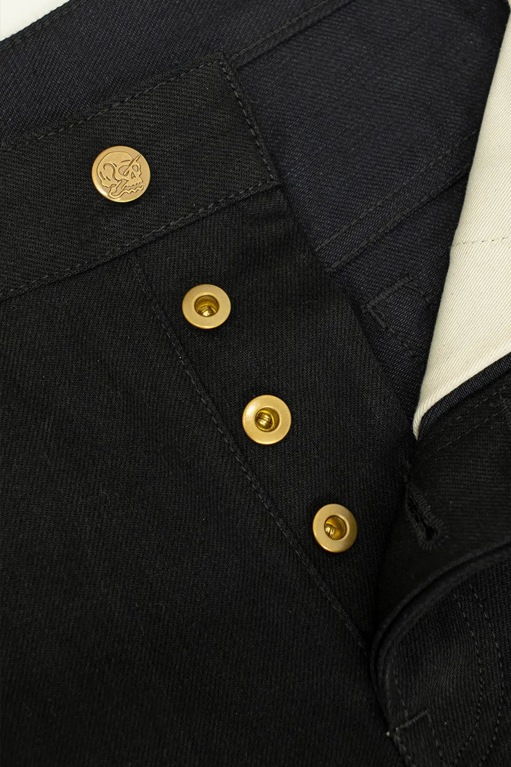 Close-up of a black denim jacket featuring four gold-tone snap buttons on the chest and a circular gold logo medallion on the pocket flap.