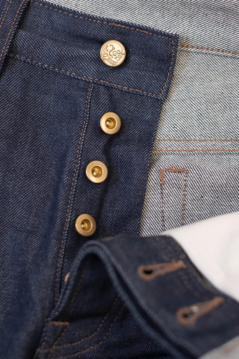 Close-up of dark blue denim jeans showing a button fly with gold-tone metal buttons and a light blue denim side panel.