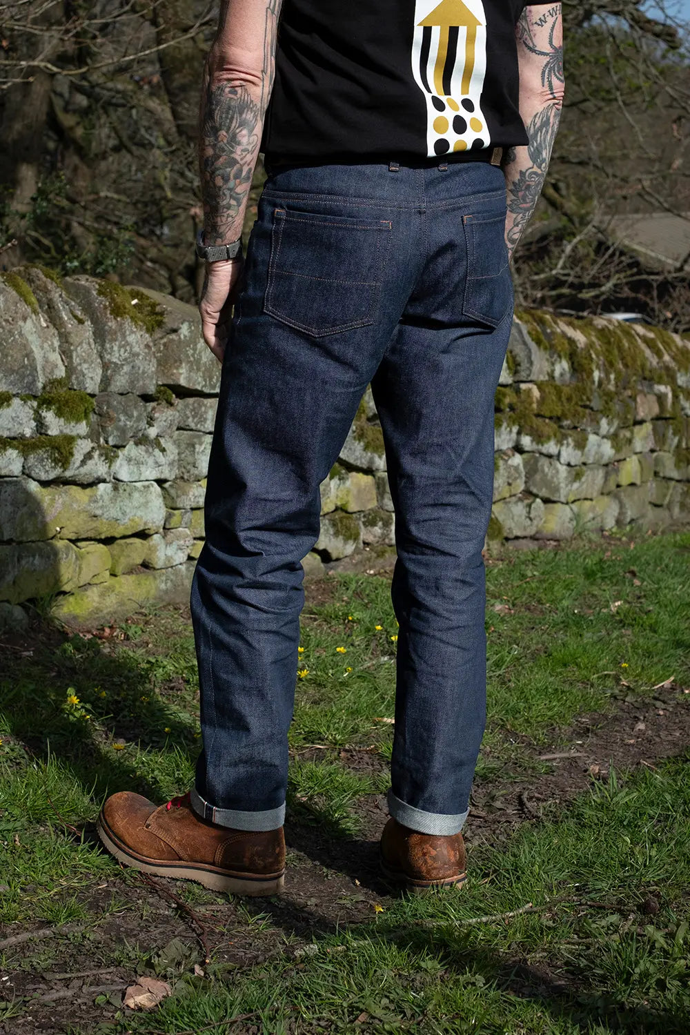 A person standing outdoors in front of a stone wall, wearing dark blue denim jeans, a black graphic t-shirt, and brown boots.