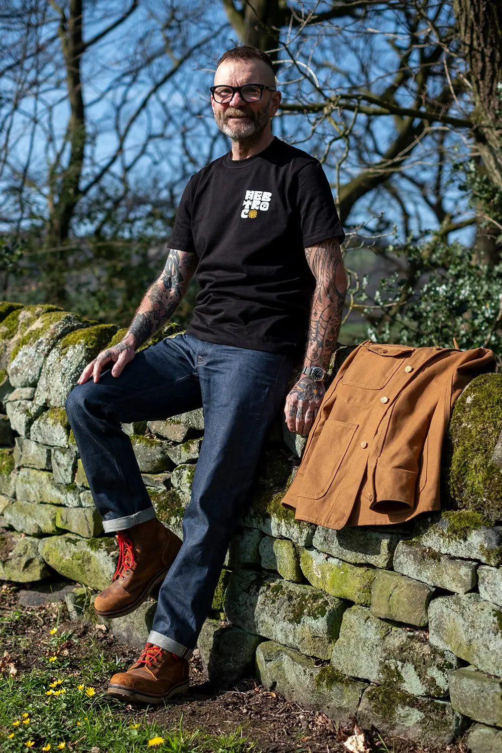 A man wearing a black graphic t-shirt and blue jeans leans against a mossy stone wall outdoors, with a brown jacket resting on the wall next to him.
