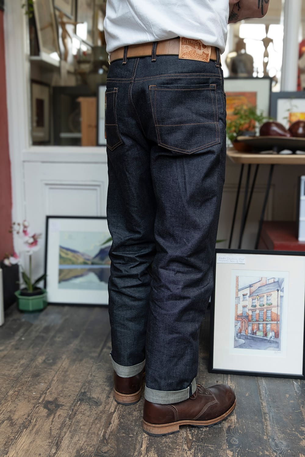 A person shown from the back wearing dark indigo denim jeans with a brown leather belt, white shirt, and brown boots in a styled indoor setting with framed art on a wooden floor.