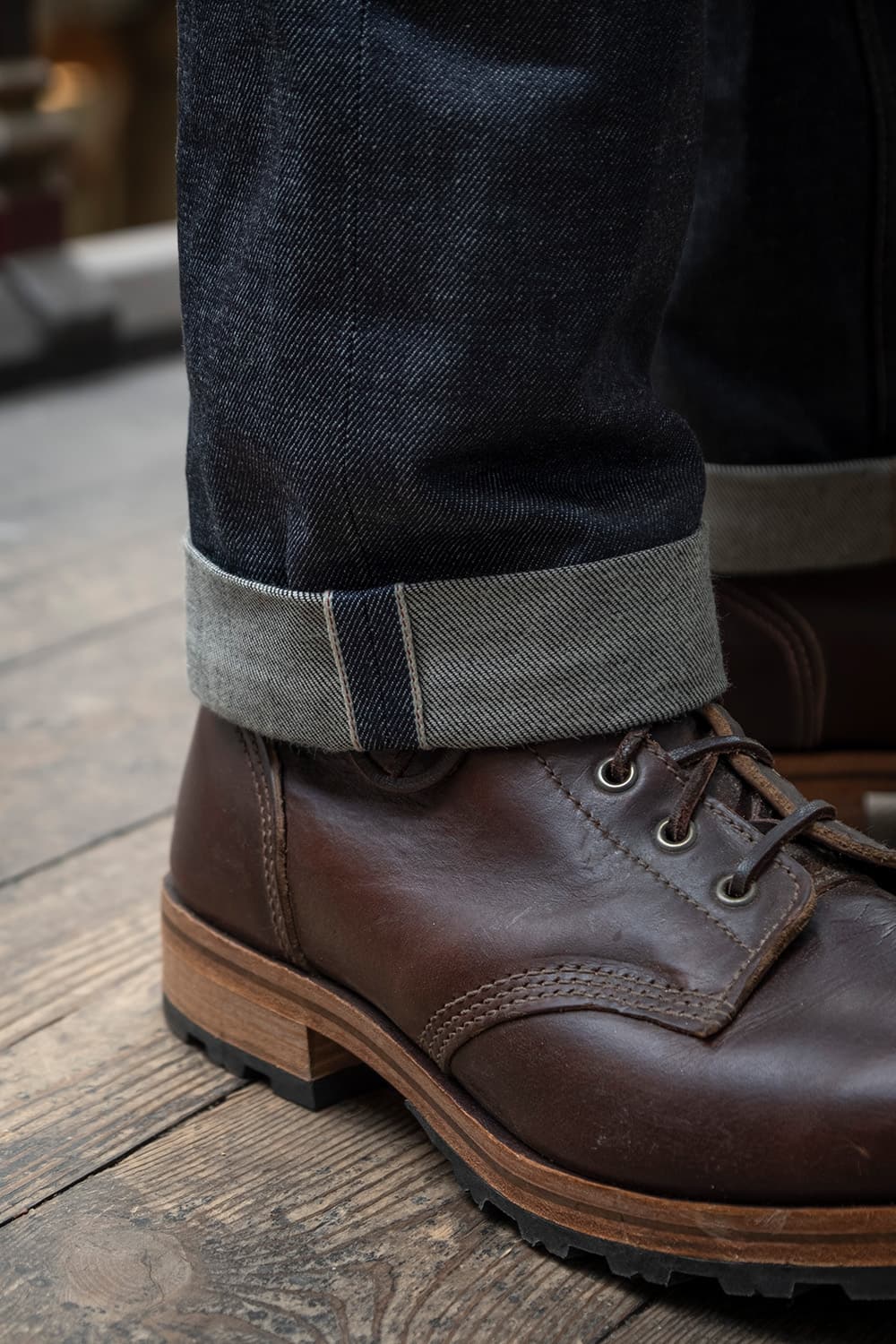 A close-up shot of a person's foot wearing a dark brown leather lace-up work boot with a cuffed dark blue denim jeans, standing on a rustic wooden plank floor.