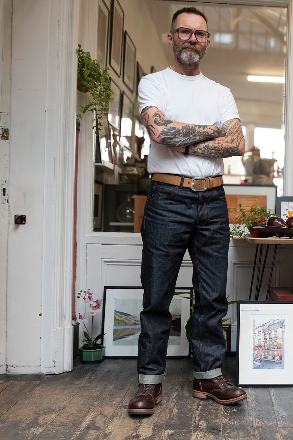 A man with glasses, a beard, and tattoos stands in a room with his arms crossed, wearing a white t-shirt, dark blue jeans, a brown belt, and brown leather boots.