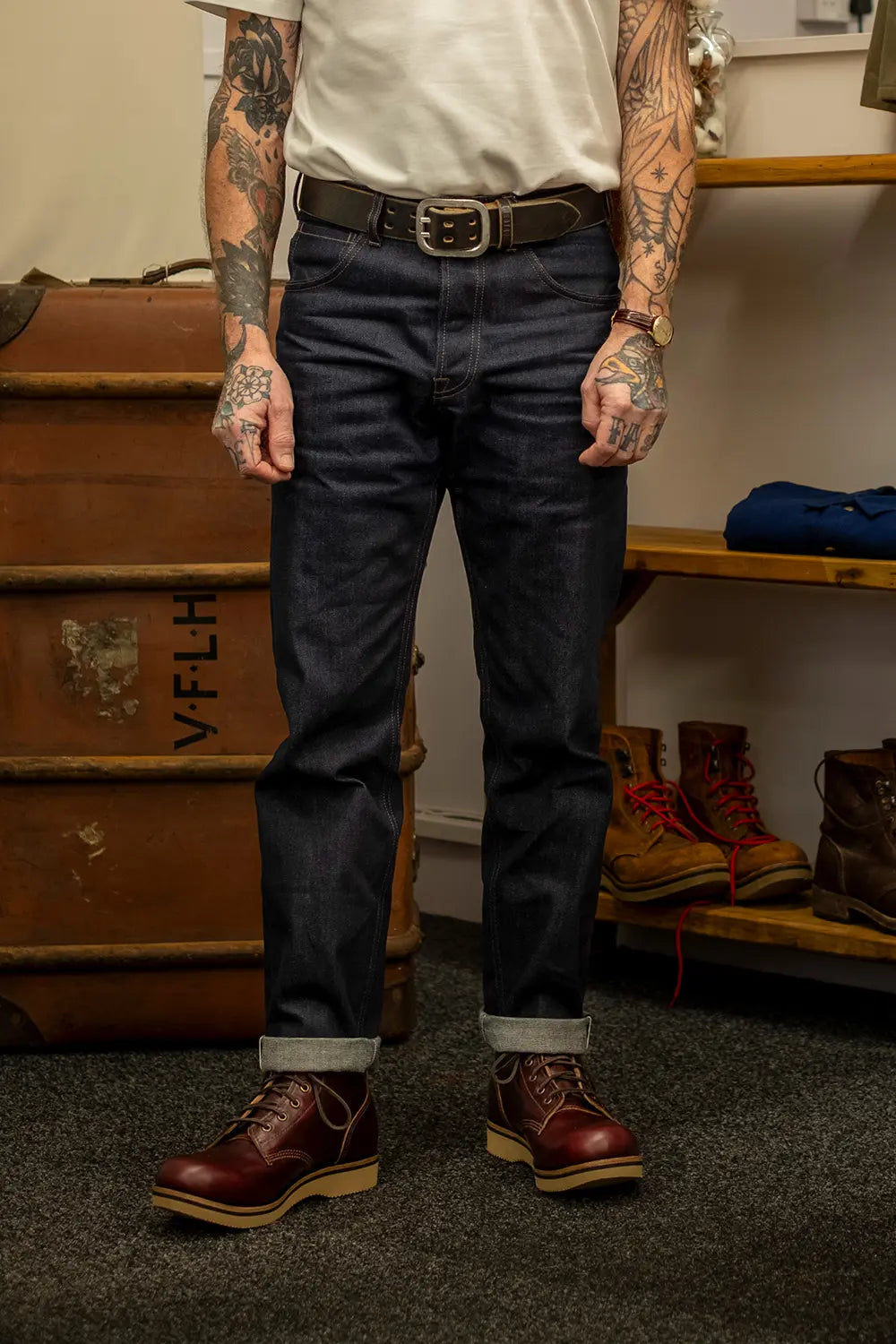 A person wearing dark indigo jeans with a black belt, styled with a white t-shirt and brown leather boots in a rustic indoor setting with vintage wooden crates and shelves.