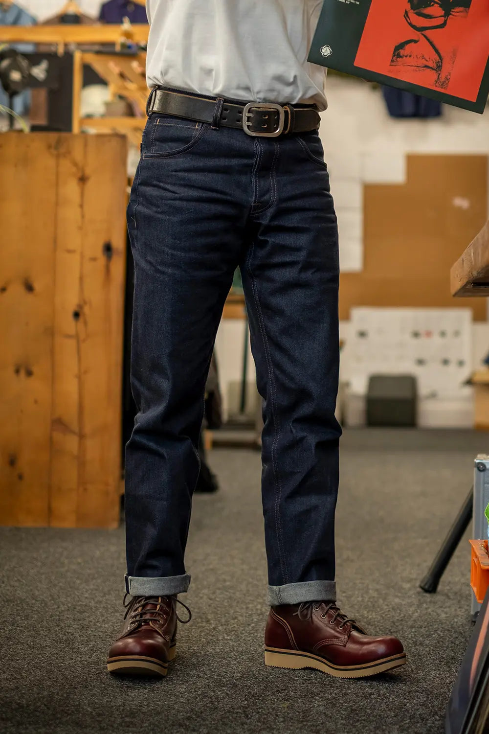 A person shown from the waist down wearing dark blue denim jeans with a black belt, paired with a white shirt and brown leather lace-up boots in an indoor workshop or studio setting.