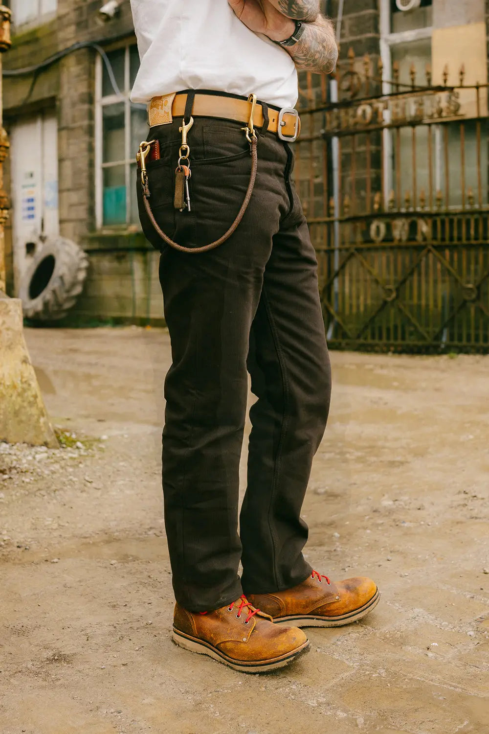 A person standing outdoors wearing dark brown work pants with a tan leather belt, a white shirt, and tan leather work boots. The photo is cropped from the chest down, showing the fit of the pants and the styling in a rustic outdoor setting.