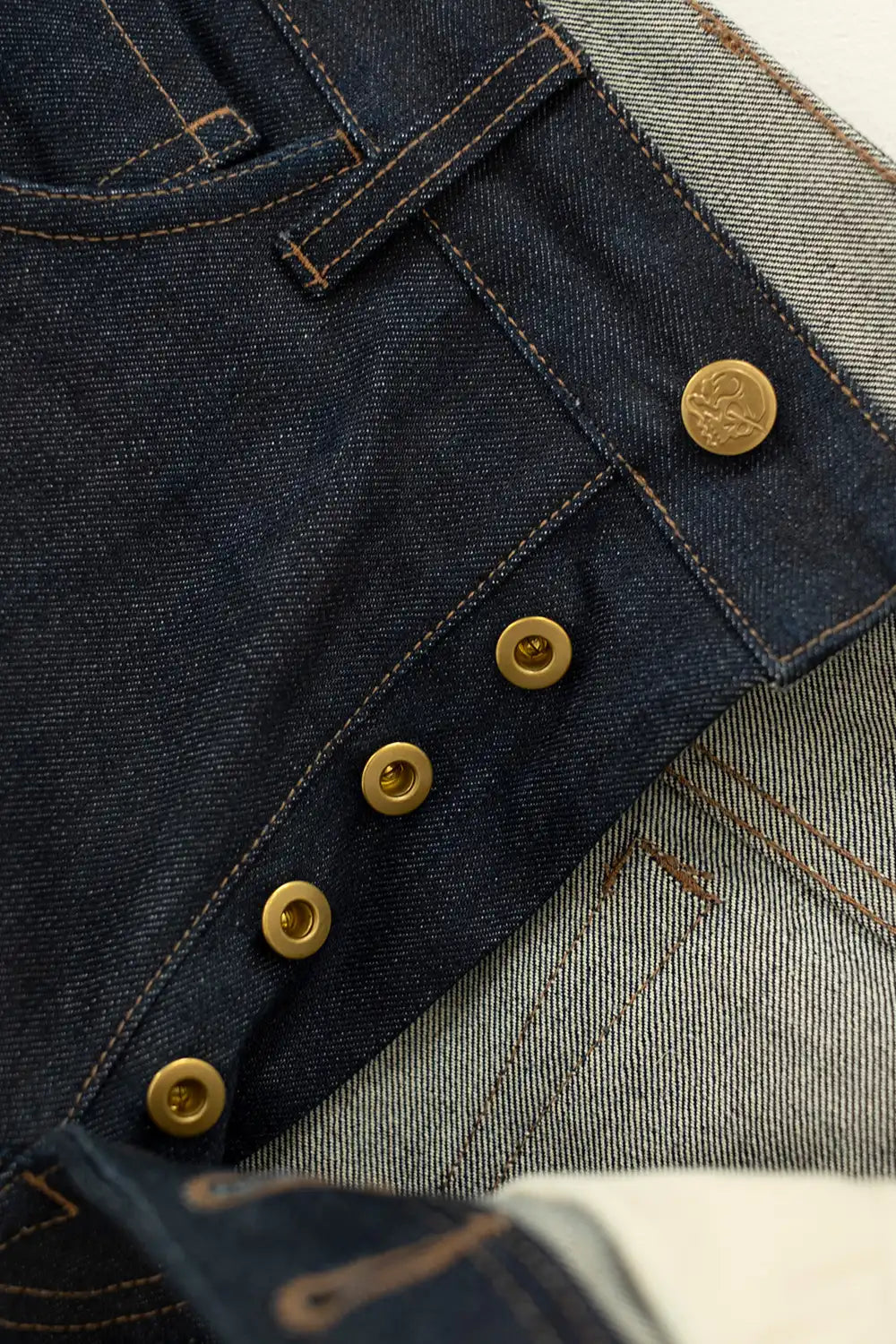 Close-up of dark indigo denim jeans showing the button fly closure with brass buttons and contrast stitching, layered over a lighter gray denim fabric.