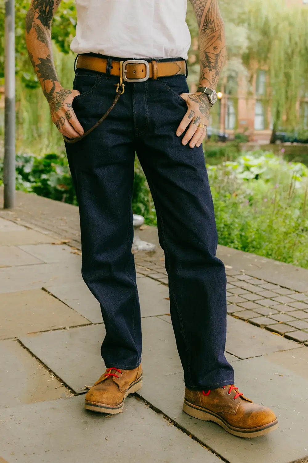 A person outdoors wearing dark navy blue jeans, a brown leather belt, a white t-shirt, and brown lace-up work boots, standing on a stone walkway with greenery in the background.