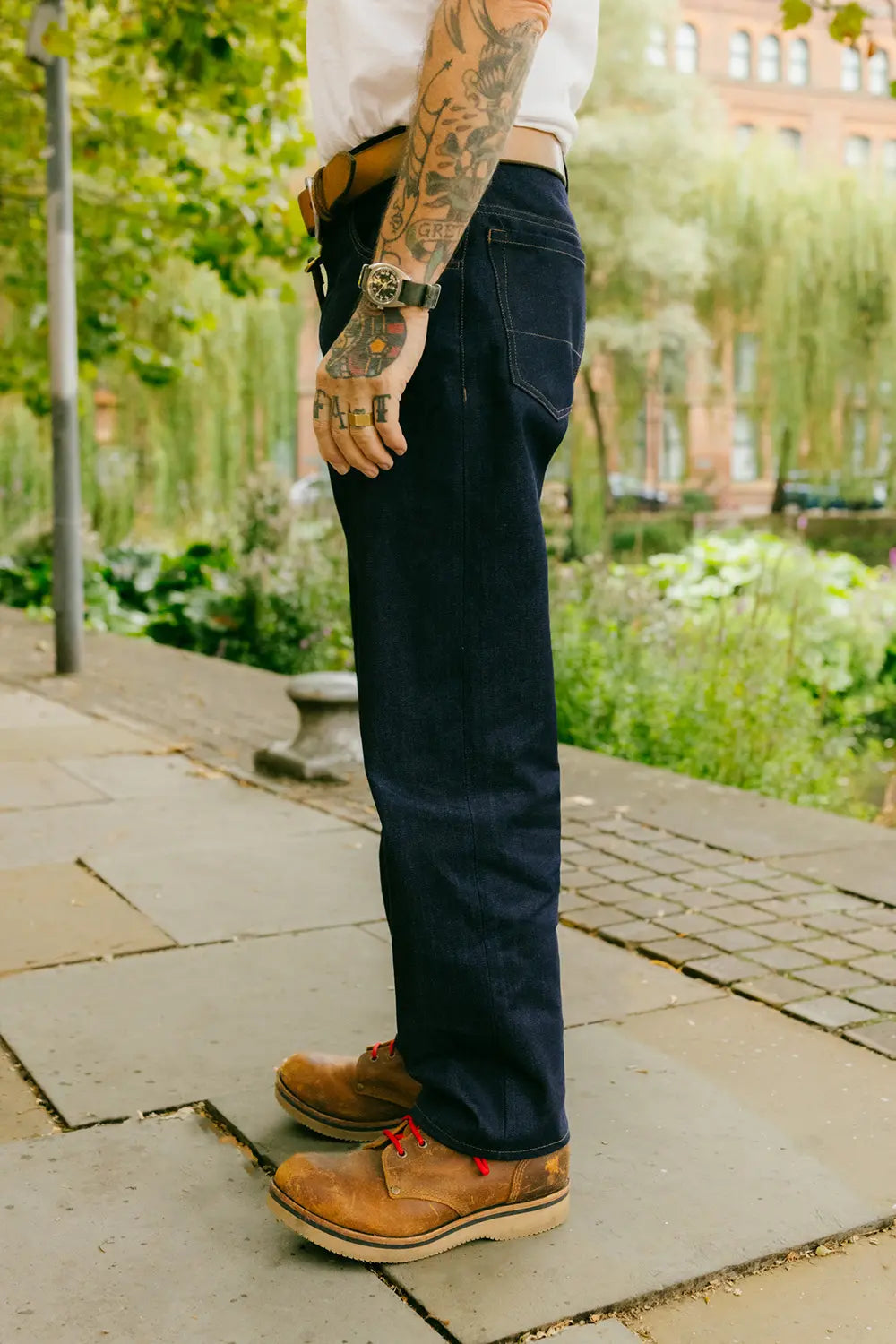 A person standing outdoors on a stone walkway wearing dark indigo jeans, a white shirt, and brown leather boots, with greenery and a building blurred in the background.