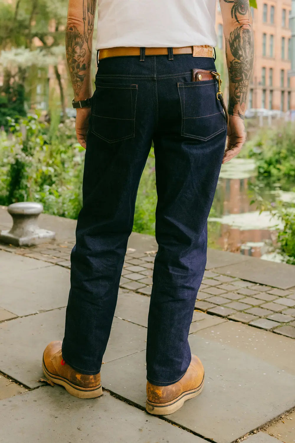 A person viewed from behind wearing dark indigo denim jeans, a white t-shirt, a tan leather belt, and brown work boots, standing outdoors on a stone walkway.