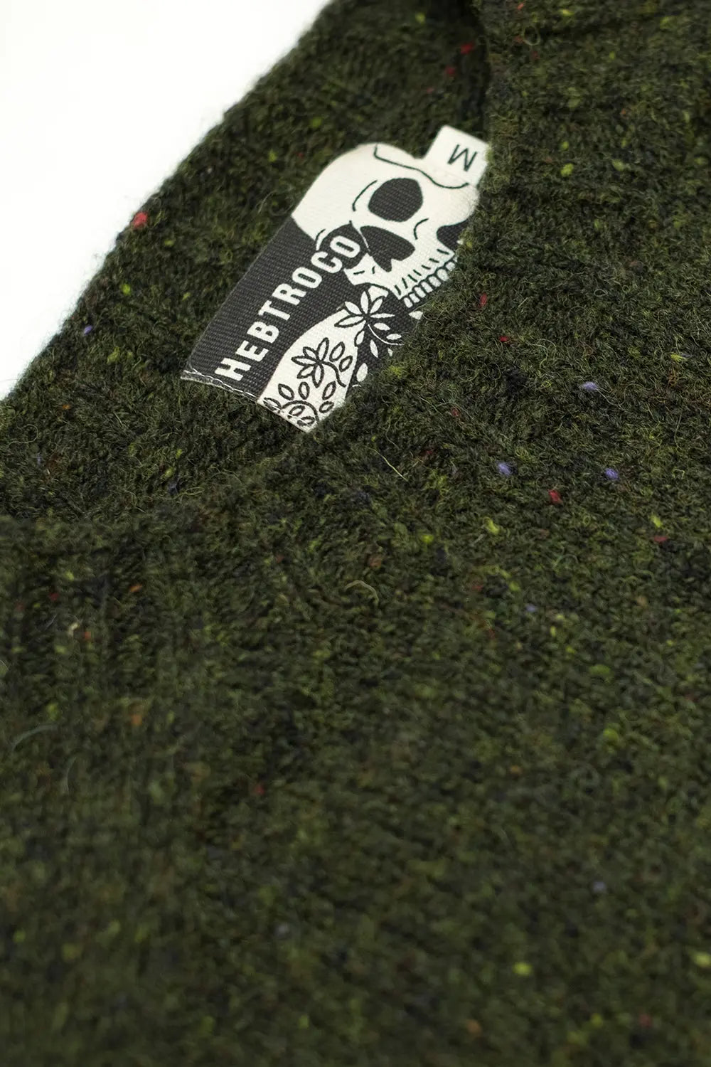 Close-up of a dark olive green textured knit fabric with tiny multicolor flecks, featuring a sewn-on brand label for 'Hebtroco' with a white skull graphic.
