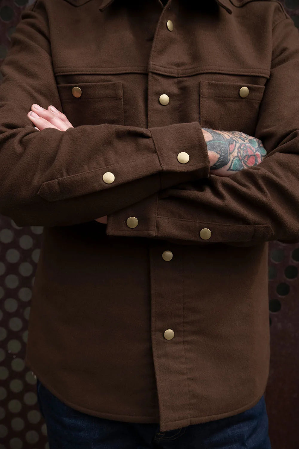 A close-up of a person wearing a dark brown moleskin iron shirt with brass-coloured snap buttons and flap chest pockets, paired with blue jeans.