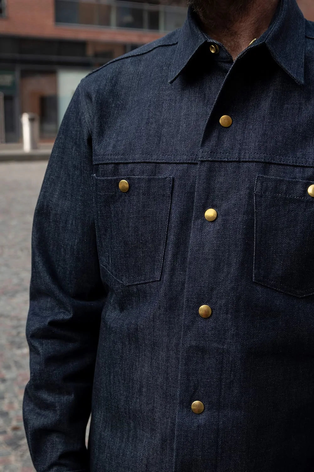 A close-up of Ed wearing a dark indigo denim ironshirt with brass buttons, set against an out-of-focus urban street background.