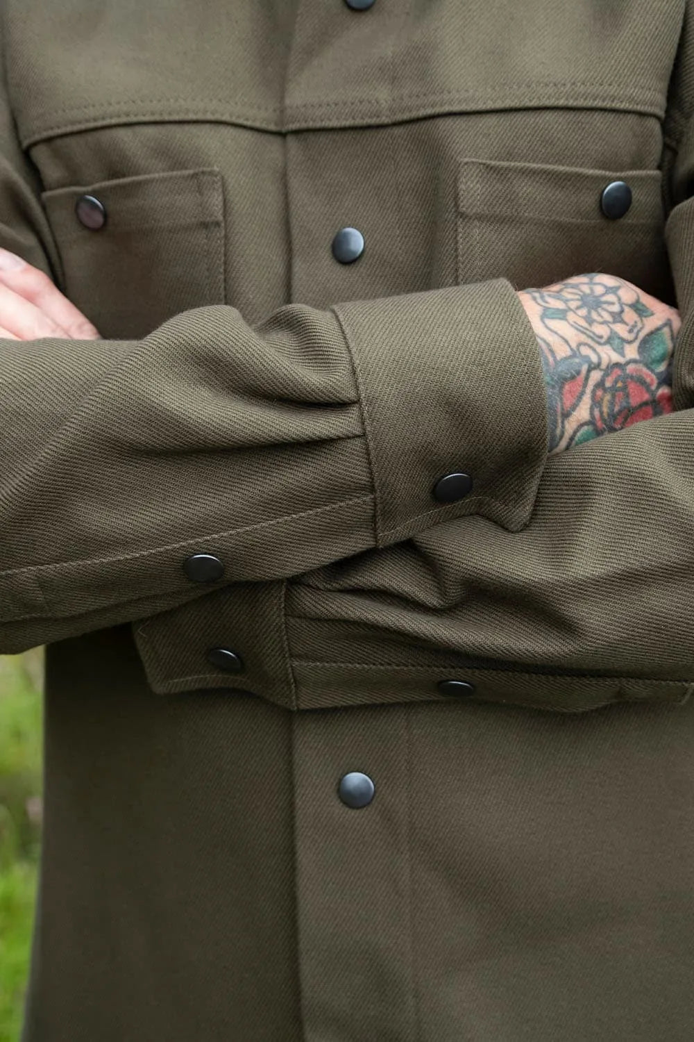 Close-up of a Ed wearing a kharki ironshirt with snap buttons and chest pockets, showing the fabric texture and a tattooed forearm.