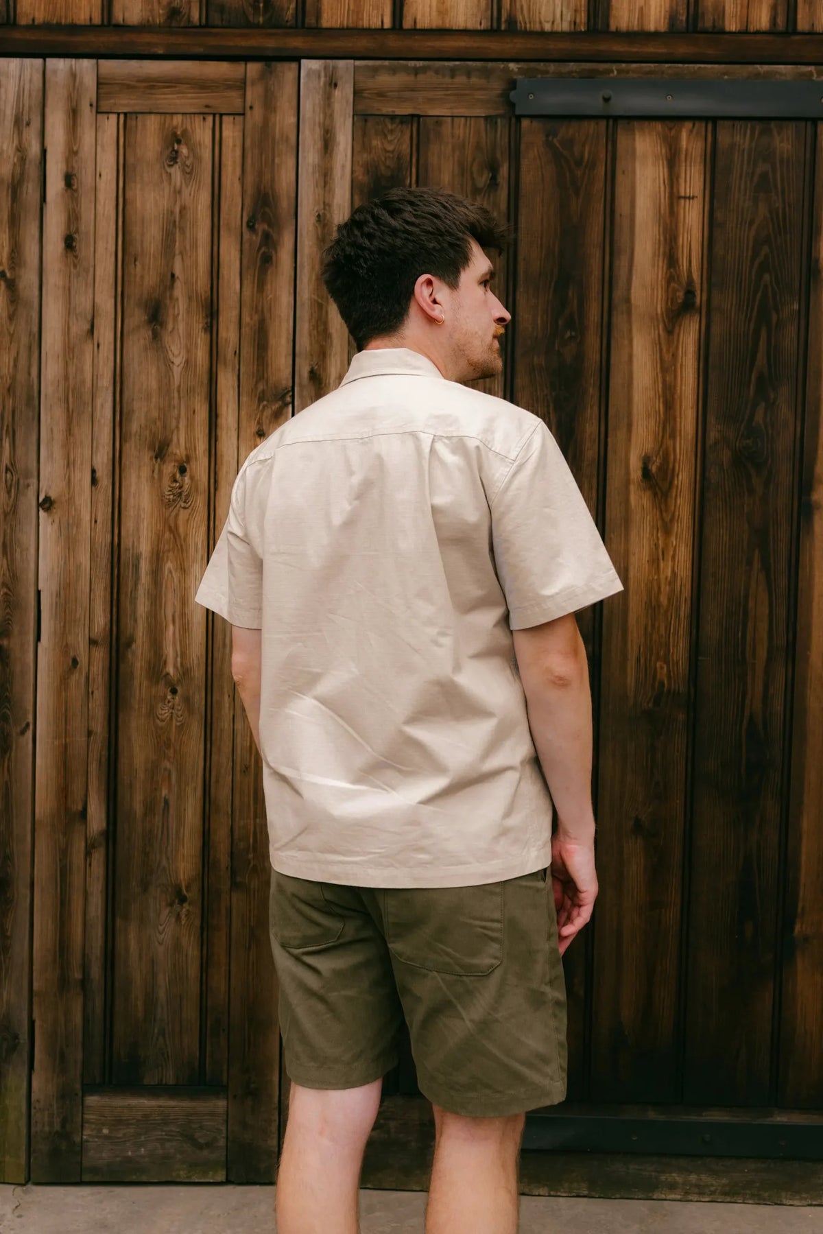 A man wearing a light beige short-sleeve button-up shirt and olive green shorts, standing in front of a rustic brown wooden plank wall.