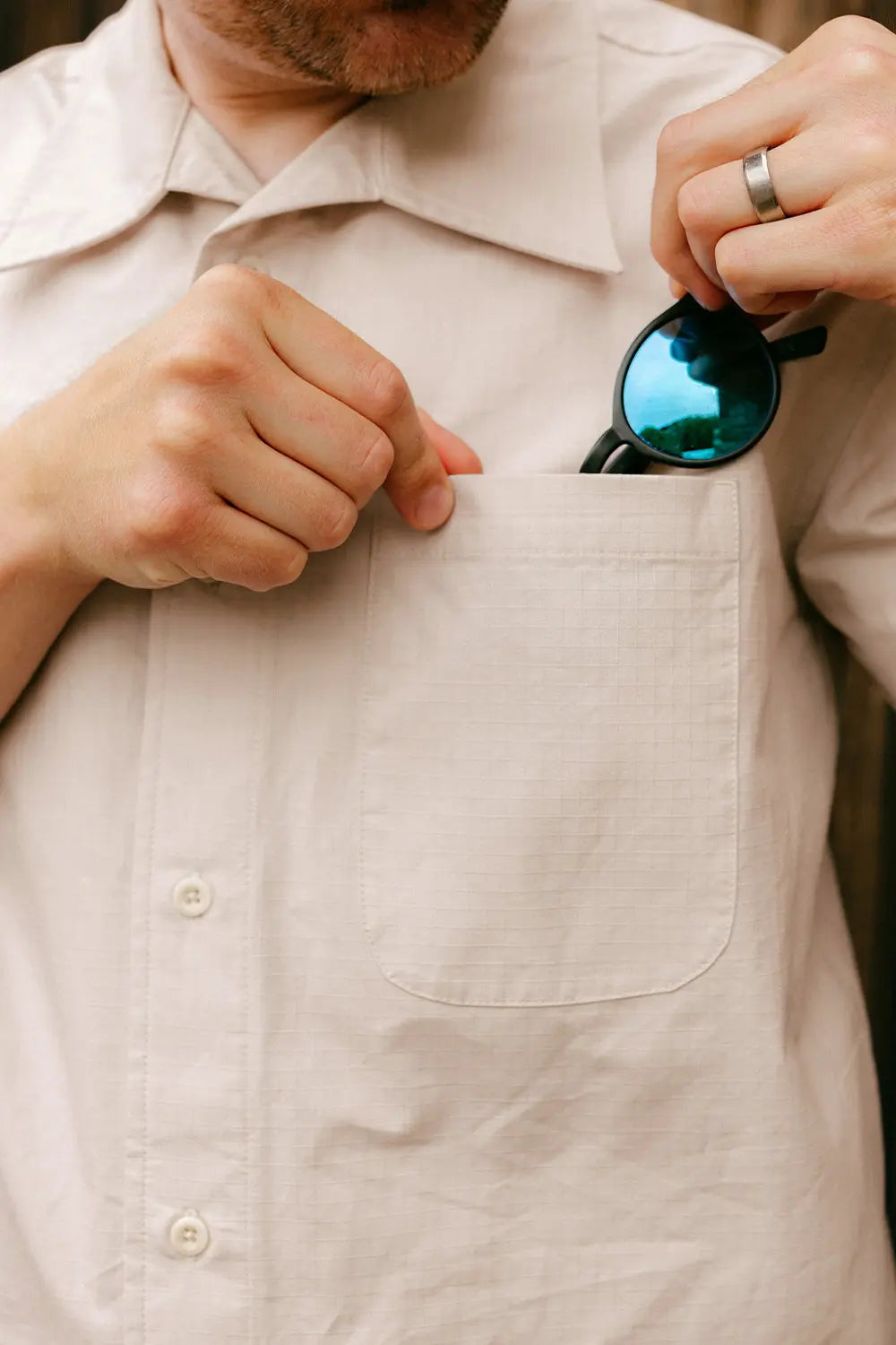 A close-up of a man wearing a light beige linen button-down shirt, holding a pair of blue mirrored sunglasses in the pocket of the shirt.
