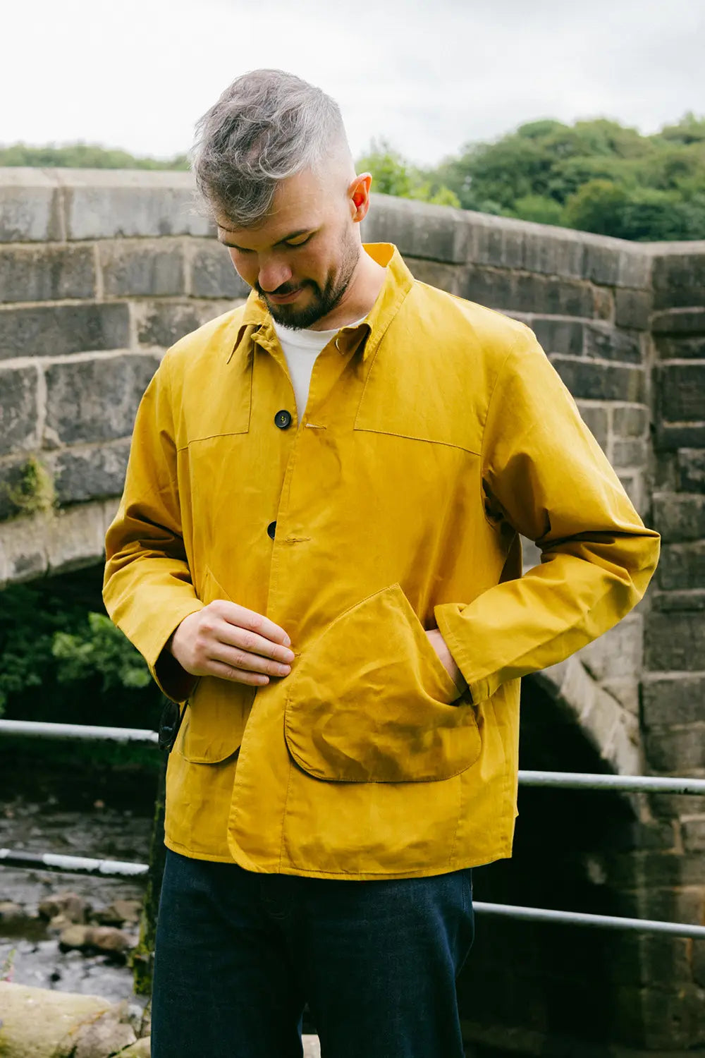 A man wearing a mustard yellow waxed cotton jacksnipe jacket over a white t-shirt and dark trousers, standing outdoors in front of a stone bridge.