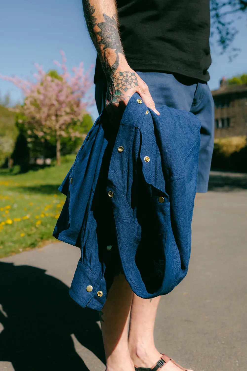 A person outdoors holding a dark blue button-up shirt in one hand, wearing a black top and blue shorts, with a sunny park background.