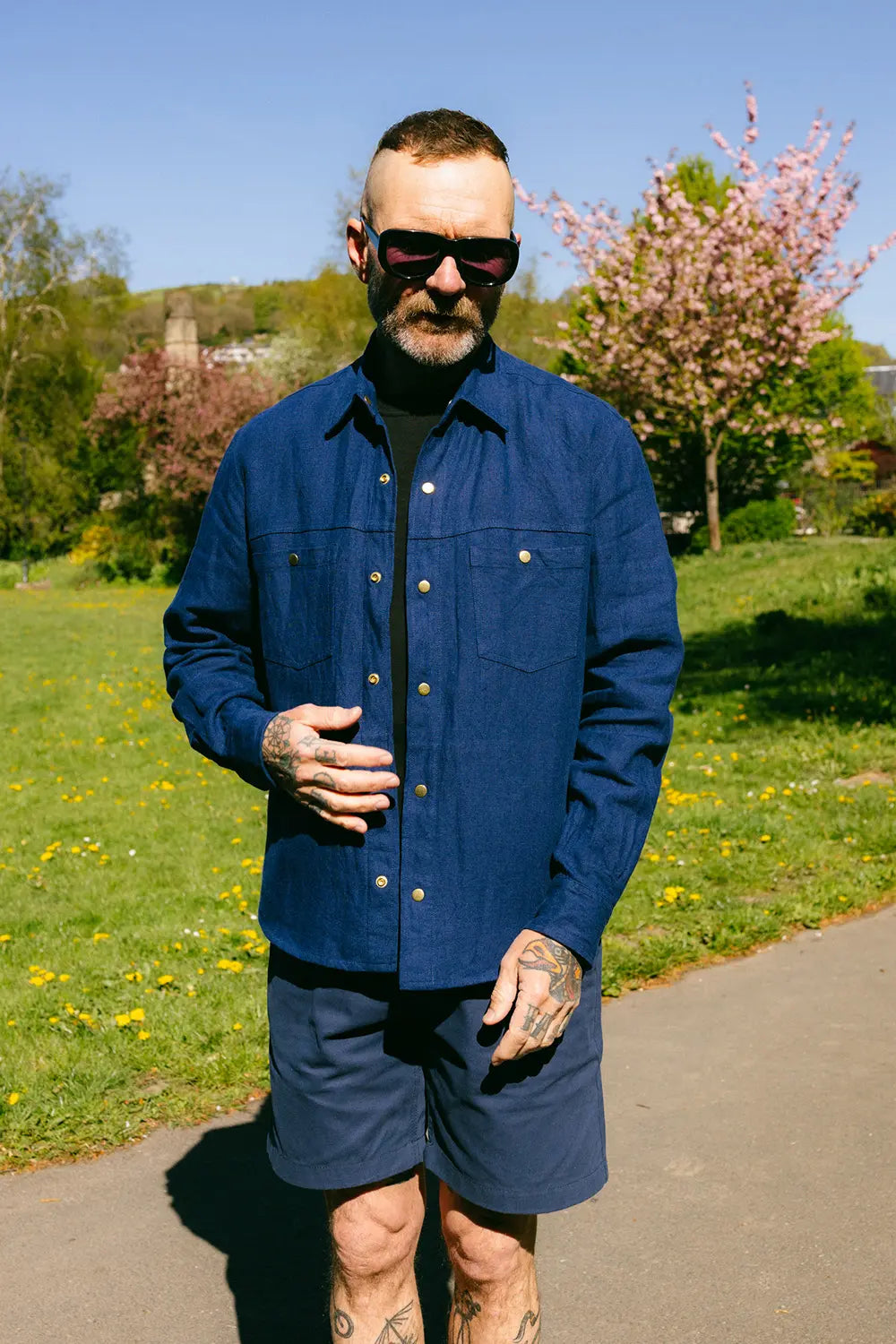 A man wearing a dark blue button-up overshirt and matching shorts, standing outdoors on a paved path with grass, trees, and a blue sky in the background.
