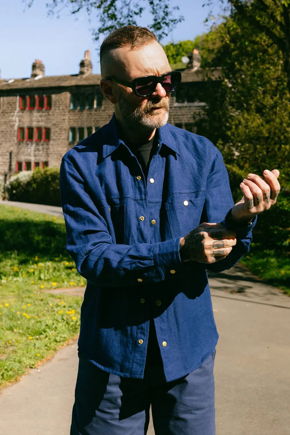 A man wearing a dark blue long-sleeve button-up shirt and matching trousers, standing outdoors on a paved path with trees and a building in the background.