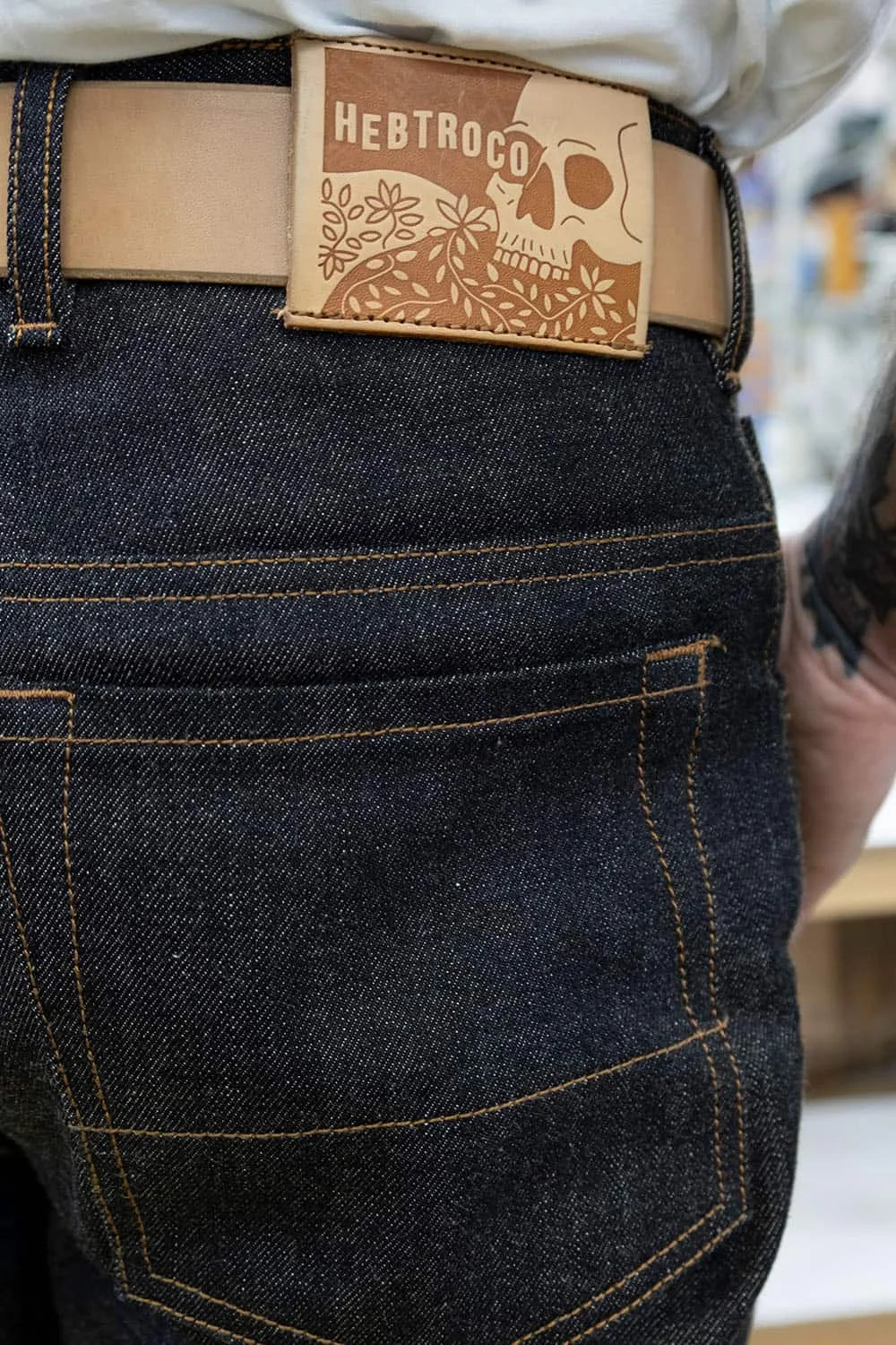 Close-up of the back waist of Ed wearing Japanese dark indigo denim jeans with contrast tan stitching, a tan leather belt, and a rectangular tan leather patch on the belt that reads 'HEBTROCO' with a skull and floral design.