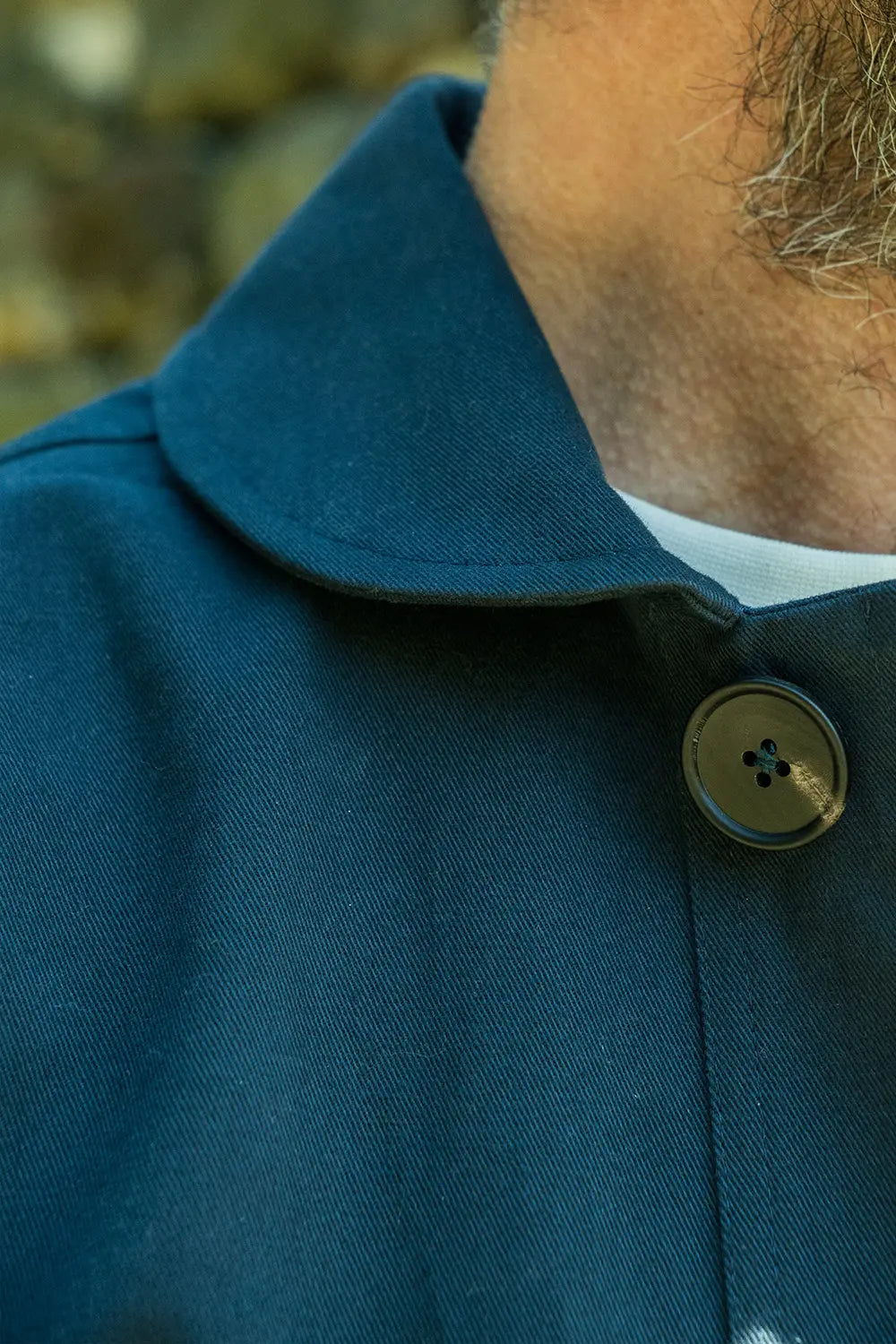 Close-up of a person wearing a dark teal-blue wool-like coat with a wide collar and a single round metallic button, with part of the neck and beard visible.