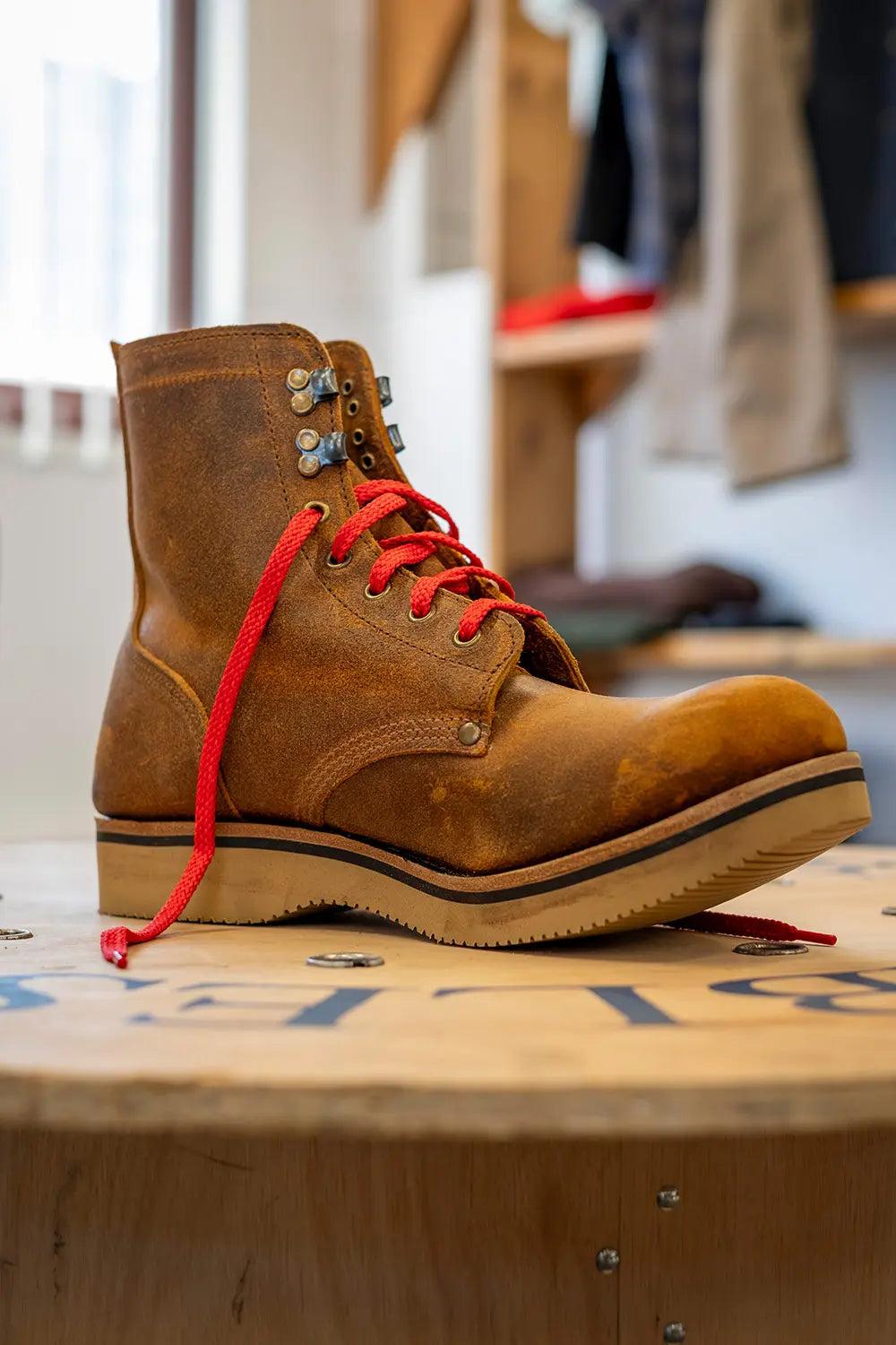 A brown leather lace-up boot with bright red laces and silver metal eyelets, resting on a wooden surface in a store setting.