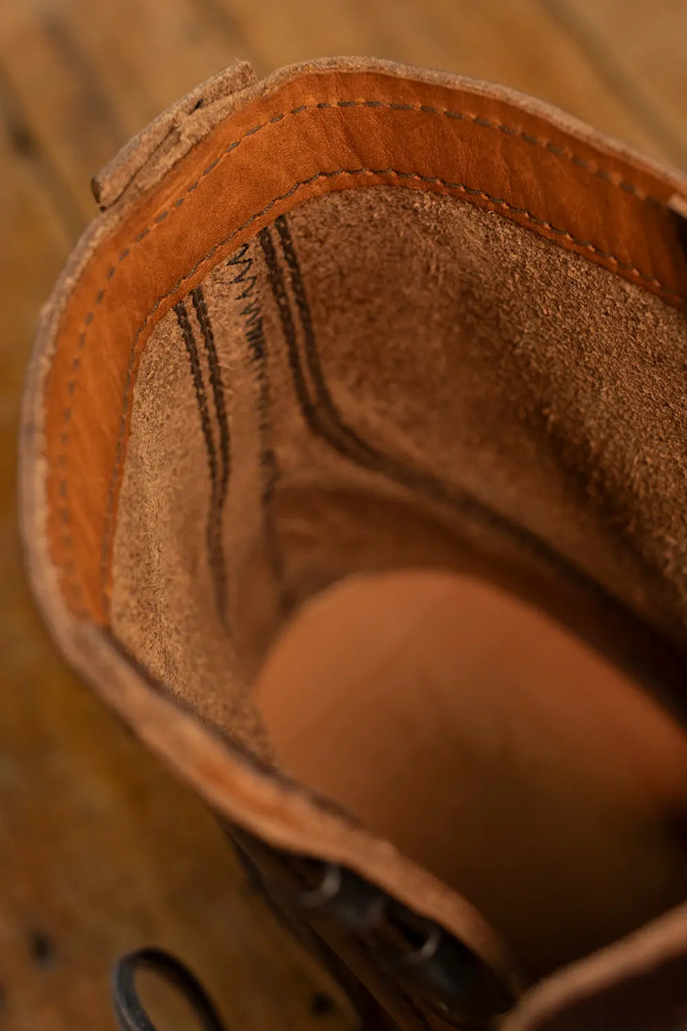 Close-up of the interior lining of a brown leather boot or shoe, showing suede-like texture, contrast stitching, and a tan footbed on a wooden surface.